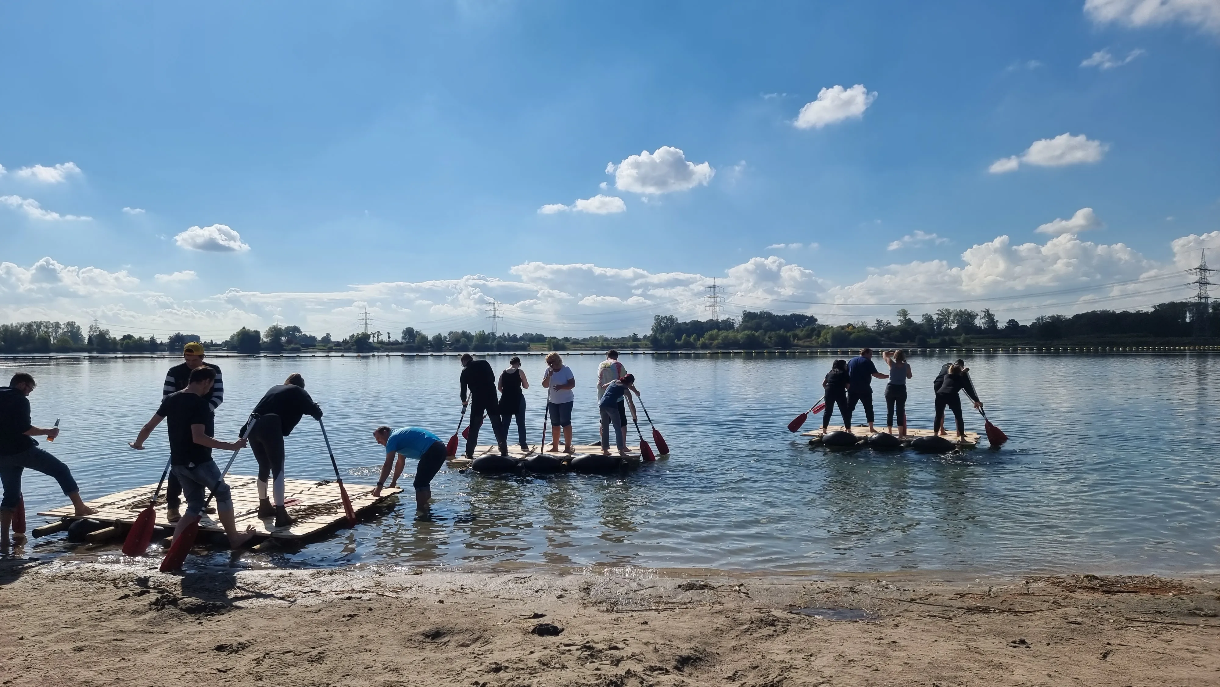 Gruppenbild an einem See, Personen stehen am Strand oder auf Stand-Up-Paddel