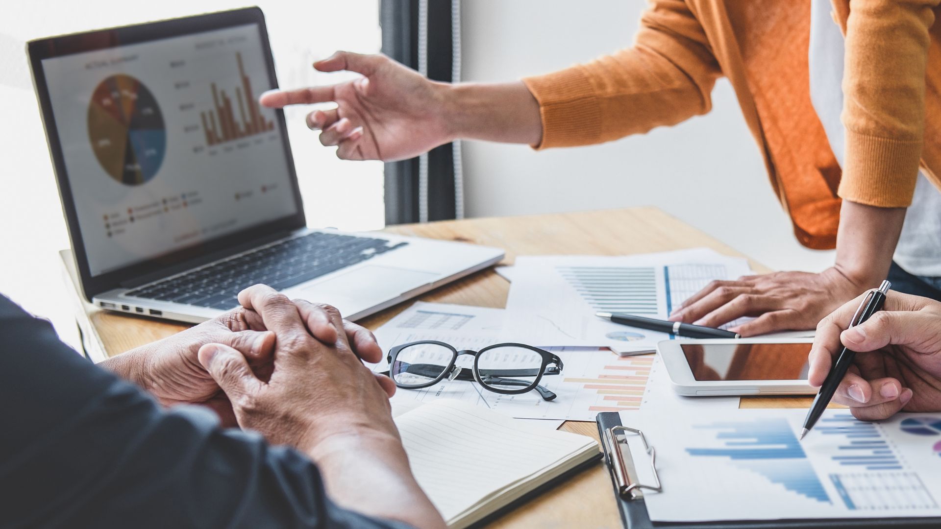 Bild: 3 Personen sitzen an einem Tisch, eine Person zeigt auf Laptop